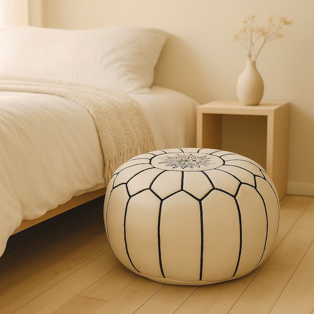 White leather pouf placed next to a cozy bed in a minimalist bedroom with light wood flooring and neutral tones.