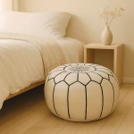 White leather pouf placed next to a cozy bed in a minimalist bedroom with light wood flooring and neutral tones.