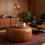 A large, wide leather floor ottoman acting as a central coffee table topped with a wooden tray and books, situated in a mid-century modern room with wood paneling and an Eames-style lounge chair.