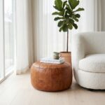A minimalist living room corner featuring a round tan leather floor ottoman used as a small coffee table with books and a mug next to a white boucle armchair and a potted fiddle-leaf fig.