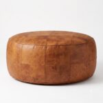 A studio product shot of a round, stitched tan leather floor ottoman with a natural grain texture, isolated against a clean white background.