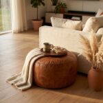 A classic tan leather floor ottoman styled with a wooden tray, a ceramic mug, and a beige knit blanket draped over the side next to a modern cream sofa.