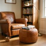 A minimalist living room corner featuring a round tan leather floor ottoman used as a small coffee table with books and a mug next to a white boucle armchair and a potted fiddle-leaf fig.