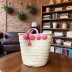 Pink pom pom straw bag with herringbone weave and tassel on wooden table in living room
