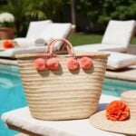 Coral pom pom straw bag with wrapped handles poolside next to straw hat and tropical flower