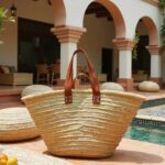 Straw tote bag beside turquoise pool, handwoven palm leaf basket with dual leather handles at Mediterranean villa