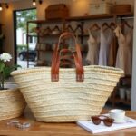 Artisan straw tote bag in boutique display, handwoven palm leaf basket with brown leather handles and white cross-stitch