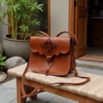 Brown leather shoulder bag displayed on woven stool with bamboo plant in natural light setting
