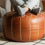 A beautiful, firm, and fully stuffed natural brown Moroccan leather pouf being used as a footrest in a sunlit, bohemian living room.