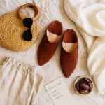 Flat-lay travel ensemble featuring rust suede Moroccan slippers, straw bag, sunglasses, linen shorts, and market-list note on cream fabric.