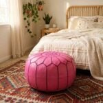 A pink leather round ottoman resting on a colorful patterned area rug at the foot of a bed in a bohemian-style bedroom with a rattan headboard and hanging plants.