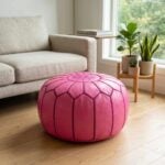 A bright pink leather round ottoman sitting on a hardwood floor in a modern living room, placed next to a beige sofa and a wooden plant stand.