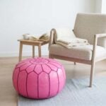 A pink leather round ottoman positioned on a light grey rug next to a comfortable beige armchair and a small wooden side table in a bright, minimalist space.