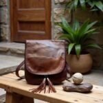 Brown leather crossbody bag with tassel accents on rustic wooden bench, outdoor courtyard setting with plants