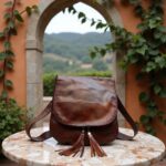 Handcrafted leather crossbody bag in brown with tassels on marble table, Italian countryside archway backdrop