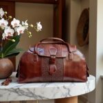 Full-grain brown leather travel bag for men on marble table with white orchid in modern entryway