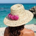 Woman wearing handwoven straw beach hat with pink pompoms at beach, wide brim sun protection Moroccan style