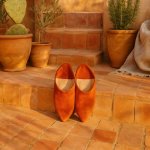 Orange suede Moroccan slippers on terracotta tiles with plants, highlighting handcrafted pointed babouche footwear