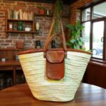 Handwoven straw shoulder bag with leather trim and front pocket resting on wooden table in rustic room