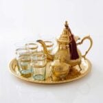Handcrafted brass tea set 9-piece with sugar bowl, featuring an engraved teapot with a burgundy tassel, six glasses with brass holders, and a matching serving tray isolated on a clean white background.
