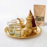 Traditional brass tea set 9-piece with sugar bowl, displayed alongside a kraft paper pouch of Moroccan green tea leaves on a bright white surface, highlighting the complete brewing experience.
