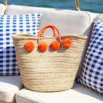 Orange pom pom straw bag with wrapped handles on boat seating with blue gingham pillows