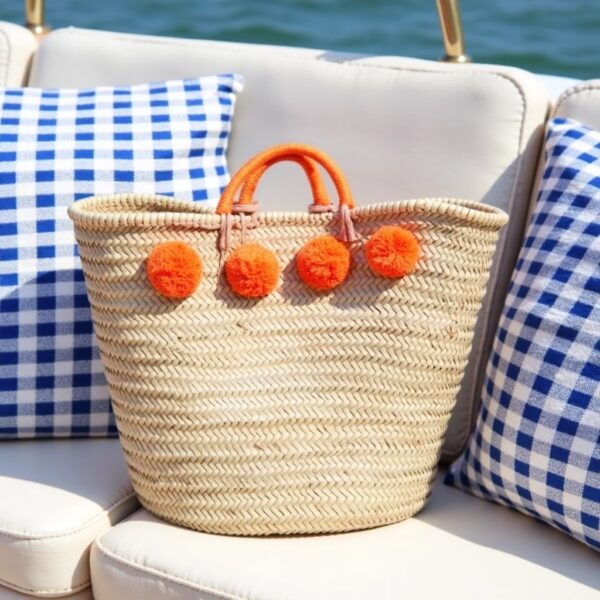 Orange pom pom straw bag with wrapped handles on boat seating with blue gingham pillows