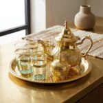 A handcrafted moroccan brass tea set including a teapot and six gold-trimmed glasses displayed on a brass-topped table with a neutral background.