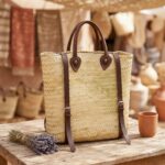 Handwoven straw backpack purse with leather straps and brass rivets displayed on a weathered wood table at a Moroccan outdoor market