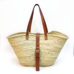 Handwoven palm leaf belted straw bag with brown leather straps and metal buckle on white background