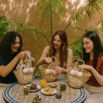 Women at table with personalized straw bag set displaying custom embroidered names and coordinating pom poms