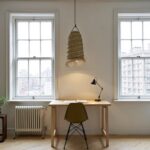 Handwoven straw lighting pendant centered over wooden desk between windows casting warm ambient glow