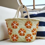 Small straw bag with orange floral design resting on striped cushion displaying artisan weaving