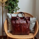 Burgundy brown leather tote bag on wicker chair showing brass buckles and contrast stitching