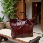 Burgundy brown leather tote bag on wooden stool showing dual buckle straps and brass hardware