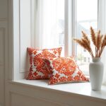 Embroidered cotton cushion pair featuring orange damask pattern on gray sofa with large window view
