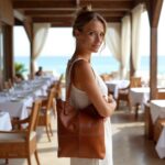 Woman wearing cognac leather hobo handbag on shoulder at beachside restaurant, showing versatile style