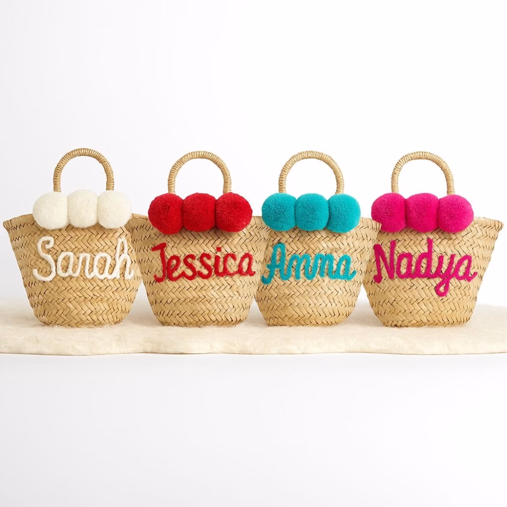 Studio lineup of four personalized mini straw bag designs, arranged in a semi-circle to show embroidered names and pom-pom colors.