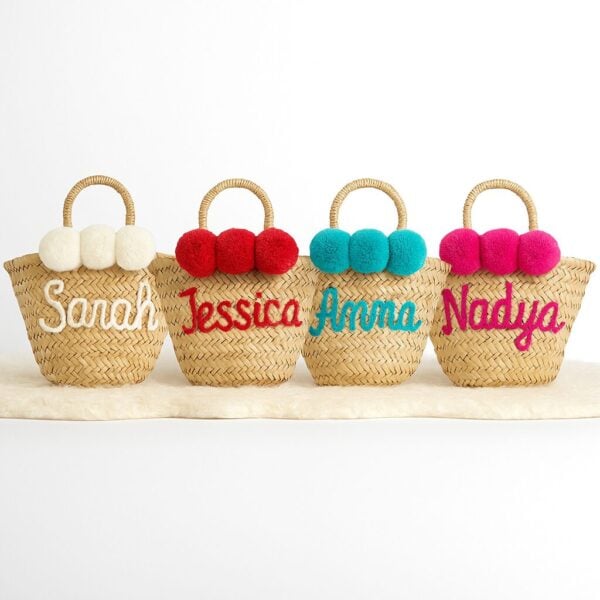 Studio lineup of four personalized mini straw bag designs, arranged in a semi-circle to show embroidered names and pom-pom colors.