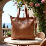 Tan leather shopper bag with studded base and dual handles on marble table, garden backdrop