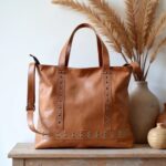 Full-grain tan leather shopper bag with crossbody strap and decorative rivets, neutral decor