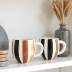 Handmade ceramic mugs with gold, black and brown vertical stripes displayed on white shelf with modern decor