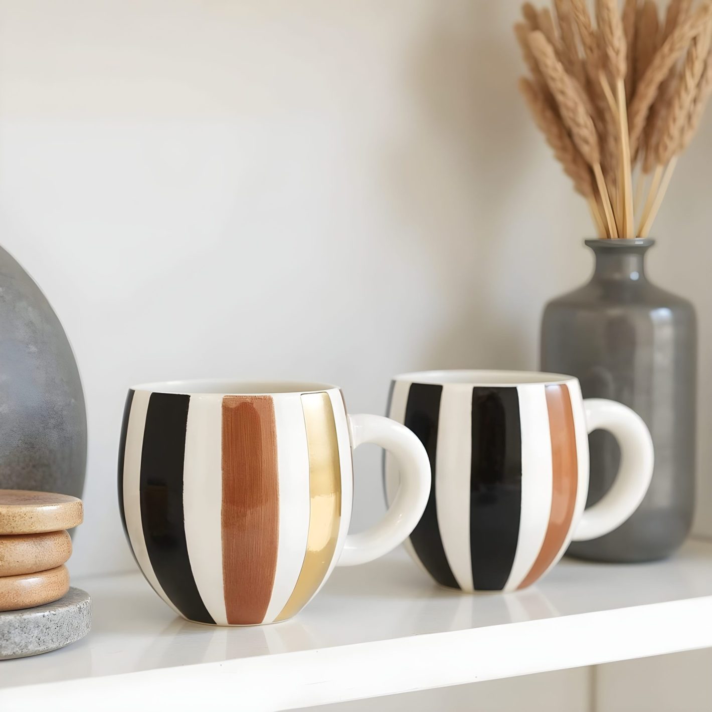 Handmade Pottery Mugs with Gold Ornaments Handmade ceramic mugs with gold, black and brown vertical stripes displayed on white shelf with modern decor