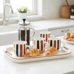 Set of four ceramic mugs with gold stripes on kitchen tray with croissants and French press coffee maker