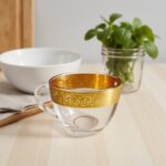 Gold scrollwork and glass handles on clear glass coffee cups resting on a rustic wood surface