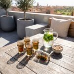 Sunlit oriental tea glasses filled with tea resting on a wooden patio table, accompanied by a pitcher of fresh mint water and sugar cubes, set against a scenic Moroccan rooftop view.