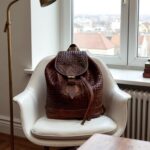 Brown basket-weave leather backpack with brass hardware on wooden bench in traditional courtyard