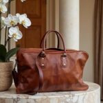 Handcrafted cognac leather weekend bag on marble table beside a potted orchid, featuring solid brass hardware