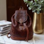 Handcrafted woven leather backpack in brown with brass buckle beside laptop and coffee mug