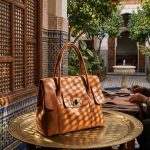 A cognac leather tote bag displayed on a brass tray in a Moroccan courtyard with an artisan working in the background.